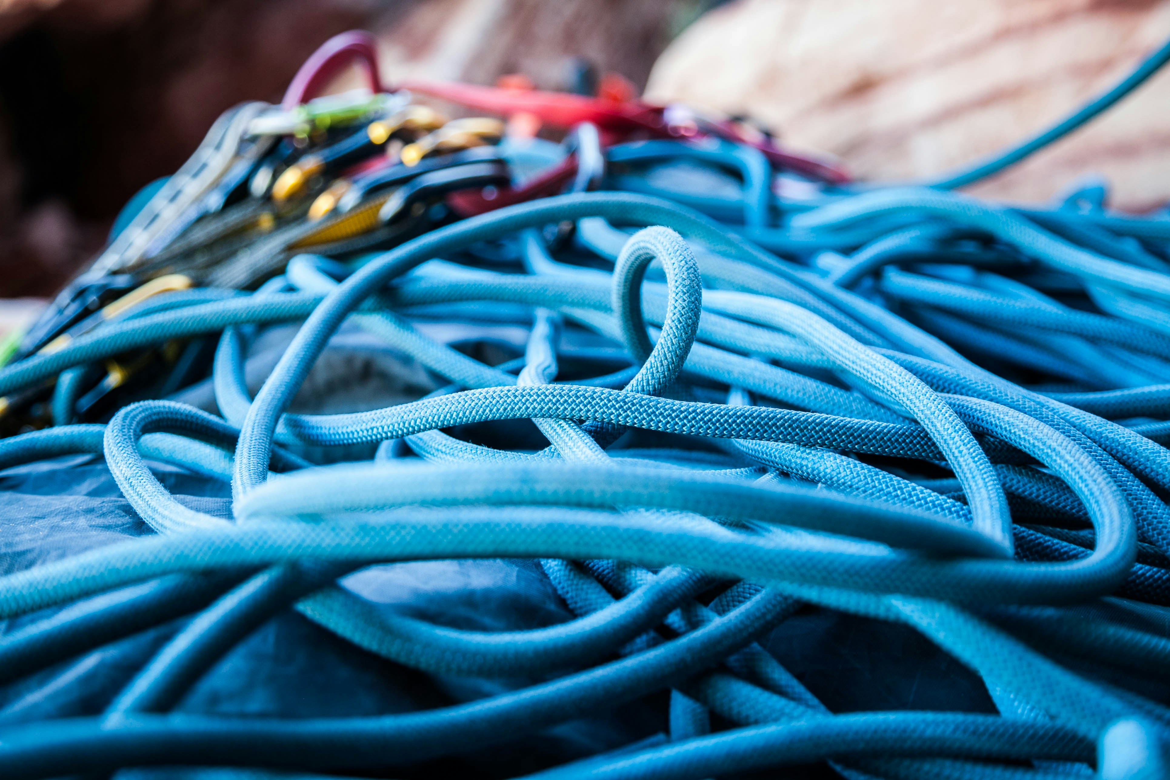 Image of a multipleblue colour ropes used for climbing, piled in a haphazard way, there are some carabiners, out of focus, in the background.