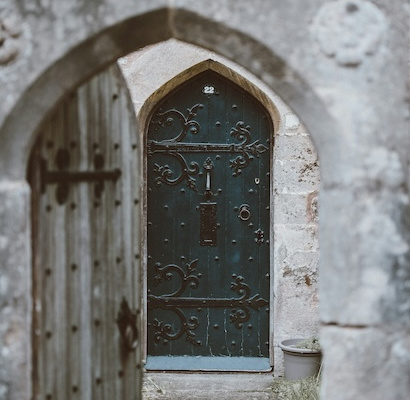 An old, heavy, grey wooden door partially open with a green door in the distance.