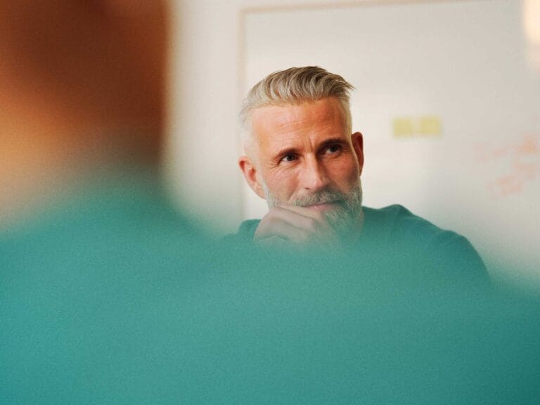 In the foreground is a persons back up close with a green shirt on. Looking over the persons shoulder is a male looking away sitting in front of a whiteboard