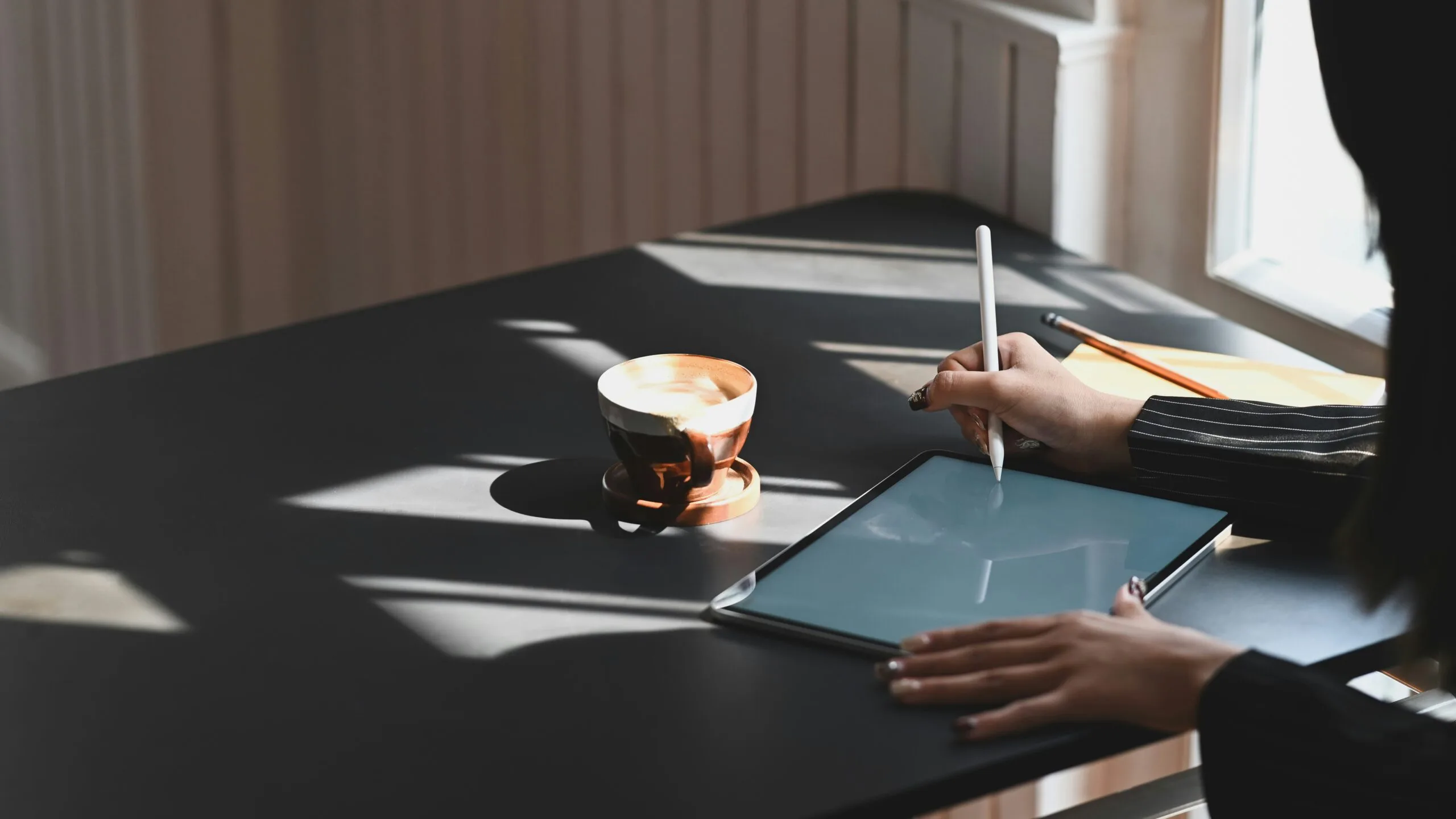 Table with a coffee mug and tablet on it. Someone is sitting at the table using a e-pen to write on the tablet