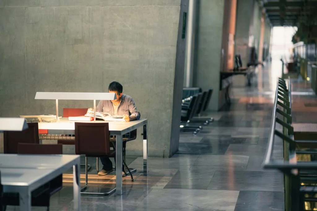 Male sitting inside a building with a open space tables for working and studying. His face is covered by a light on the desk. He is reading a book