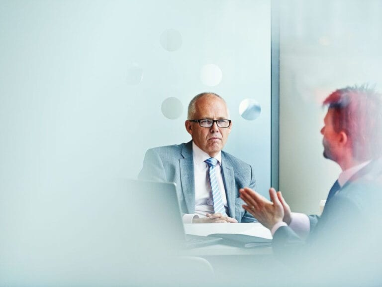 Two men sitting across from eachother, looks like the office of the male we can see. He is an older man looking at the other man who we can only see the side of his face who is talking