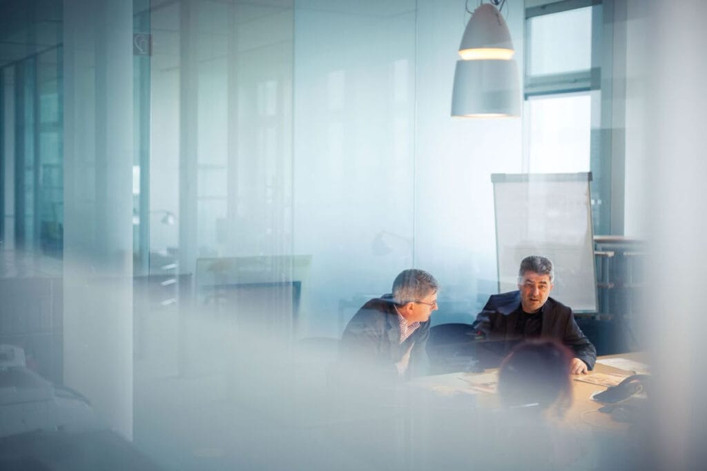 People sitting in a office around a table talking to eachother. There are 2 males we can see and the back of a woman