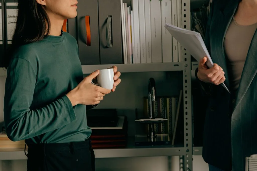 2 people talking in front shelving with books and folders on the shelf. You can't see their faces but one is a woman holding a mug in a green top on the left and another person on the right holding a stack of papers