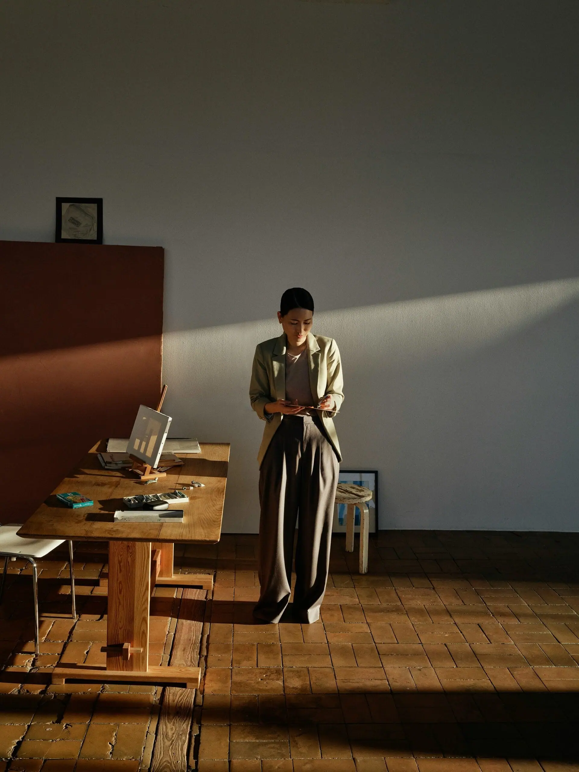 Woman standing in art studio drawing on something in her hands. Standing next to a table with tools and a small easel. Light shining in through a window on the right giving lots of shadows but also highlighting things in the room